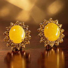 Load image into Gallery viewer, A pair of gold-toned stud earrings with oval yellow gemstones, photographed close up on a reflective dark brown surface with a warm blurred background.