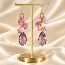 Load image into Gallery viewer, A pair of dangling earrings displayed on a gold T-stand against a draped champagne satin background. The earrings feature gold-tone metal with clusters of pink faceted beads, pearl-like white spheres, and large teardrop-shaped pink gemstones.