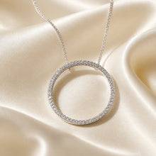 Load image into Gallery viewer, A sterling silver-toned open circle pendant necklace paved with small clear Austrian crystal stones, hanging from a delicate chain against draped champagne satin fabric.