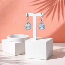 Load image into Gallery viewer, Pair of silver earrings with large clear gemstones on a white stand against a peach background