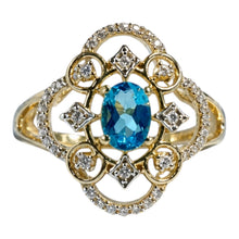 Load image into Gallery viewer, A yellow gold ring featuring a central oval-cut blue gemstone surrounded by a decorative floral cluster of small clear stones, set against a plain white background.