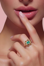 Load image into Gallery viewer, A close-up lifestyle shot of a model's hand and lips, showcasing a gold flower ring with a green emerald center and small clear accent stones.