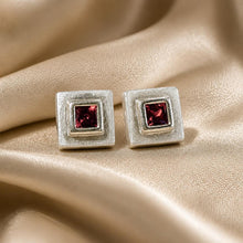 Load image into Gallery viewer, A pair of square stud earrings with textured silver-toned metal and a central square-cut deep red garnet gemstone, displayed on draped champagne-colored satin fabric.