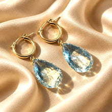 Load image into Gallery viewer, A pair of gold hoop earrings with large faceted light green amethyst teardrop gemstones, displayed on a draped beige satin fabric with warm sunlight and sparkles.