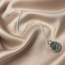 Load image into Gallery viewer, A silver chain necklace with a round textured black and silver druzy pendant, displayed on draped champagne satin fabric.