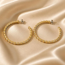 Load image into Gallery viewer, A pair of gold hoop earrings with rectangular clear stones, displayed on a draped beige satin fabric background.