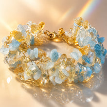 Load image into Gallery viewer, A gold-toned crystal bracelet made of mixed natural stones, including light blue, clear, and golden yellow pieces, displayed on a reflective beige surface with warm sunlight and a soft rainbow reflection.