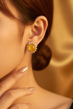 Load image into Gallery viewer, A close-up lifestyle shot of a woman wearing a gold-toned flower-shaped stud earring with a central yellow gemstone and small clear accent stones, set against a warm, draped gold fabric background.