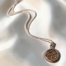 Load image into Gallery viewer, A silver chain necklace with a round, textured bronze drusy pendant, displayed on a shimmering champagne satin fabric background.