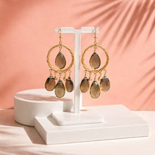 Load image into Gallery viewer, Gold earrings with teardrop-shaped stones on a white stand against a pink background