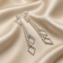 Load image into Gallery viewer, A pair of sterling silver geometric dangle earrings with open diamond-shaped charms and chain links, displayed on a draped champagne satin fabric background.