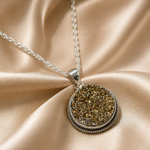 Load image into Gallery viewer, A silver-toned necklace with a round drusy gold gemstone textured pendant, displayed on draped champagne satin fabric.
