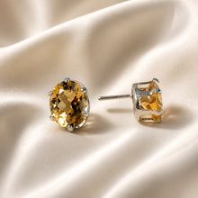 Load image into Gallery viewer, A pair of oval-cut yellow citrine gemstone stud earrings in silver prong settings, displayed on a draped cream satin fabric.
