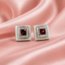 Load image into Gallery viewer, A pair of square stud earrings with textured silver-toned metal frames and a central square-cut dark red gemstone, displayed on a draped pink satin fabric background.