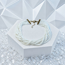 Load image into Gallery viewer, A multi-strand white beaded necklace with a gold-toned clasp and extender chain, displayed on a white hexagonal stand over clear rhinestones against a white geometric background.