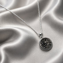 Load image into Gallery viewer, A silver chain necklace with a round bezel-set black druzy stone pendant, displayed on draped silver satin fabric.