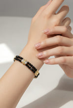Load image into Gallery viewer, A close-up of a person's wrist wearing a double-strand bracelet with black faceted beads, a gold-toned rectangular charm, and a central clear crystal cluster accent.
