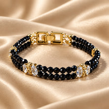 Load image into Gallery viewer, A double-strand beaded bracelet with black faceted beads, gold-toned metal links, and clear crystal rhinestone accents, displayed on a draped beige satin fabric.
