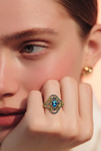 Load image into Gallery viewer, A close-up lifestyle shot of a model wearing an ornate gold ring with a central blue gemstone and surrounding green and yellow accents, resting her chin on her hand.

