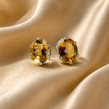 Load image into Gallery viewer, A pair of oval stud earrings with faceted yellow-gold citrine gemstones in silver prong settings, displayed on draped champagne-colored satin fabric.
