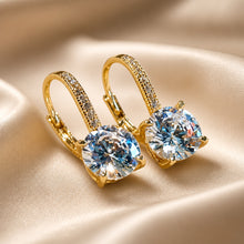 Load image into Gallery viewer, A pair of gold lever back earrings with l8 mm round clear Austrian crystal and small clear stones along the upper band, displayed on draped champagne satin fabric.
