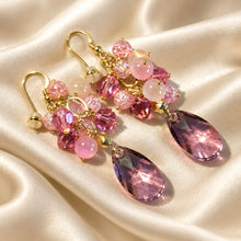 Load image into Gallery viewer, A pair of gold-tone dangle earrings with pink faceted beads, clear crystal-like spheres, and large teardrop-shaped pink gemstones, displayed on a cream satin fabric background.
