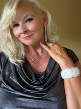 Load image into Gallery viewer, A close-up portrait of a blonde woman wearing a black cowl-neck top, layered silver necklaces, a silver drop earring, a silver ring, and a white lace cuff bracelet, set against a plain beige background.
