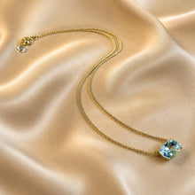 Load image into Gallery viewer, A delicate gold chain necklace with a cushion-cut clear Austrian crystal  pendant, displayed on a draped beige satin fabric background.
