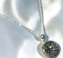 Load image into Gallery viewer, A silver necklace with a round druzy agate gemstone pendant featuring a textured, glittery black and silver druzy stone, displayed on a shiny white satin background.
