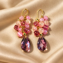 Load image into Gallery viewer, A pair of gold-tone drop earrings adorned with pink and purple faceted crystals, displayed on a draped beige satin fabric.
