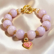 Load image into Gallery viewer, A beaded bracelet with pale pink round beads, gold-tone spacer details, a gold toggle clasp, and a dangling pink heart charm with glittery gold edges, displayed on a satin fabric background.
