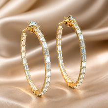 Load image into Gallery viewer, A pair of gold hoop earrings adorned with clear baguette and round-cut crystals, displayed on a draped champagne satin fabric background.
