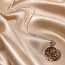 Load image into Gallery viewer, A silver-toned gemstone drusy necklace with a round textured pendant, displayed on draped champagne satin fabric.
