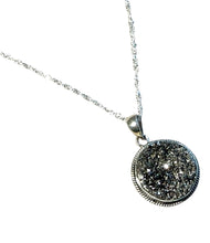 Load image into Gallery viewer, A silver-toned necklace featuring a round pendant with a textured, glittery dark gray and silver druzy stone center, set in a silver bezel with a twisted chain.
