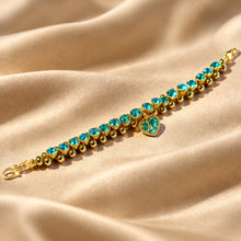 Load image into Gallery viewer, A gold-plated tennis bracelet adorned with round turquoise-blue crystal and a matching heart-shaped pendant, displayed on draped champagne-colored satin fabric.

