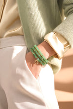 Load image into Gallery viewer, A close-up of a person's wrist wearing multiple green beaded bracelets with gold-toned metal accents, styled with a cream satin shirt, a sage green knit cardigan, and cream trousers.
