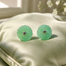 Load image into Gallery viewer, A pair of round, flower-shaped light green jadeite stud earrings with a small silver-toned four-petal charm in the center, displayed on a cream satin cushion.
