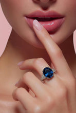 Load image into Gallery viewer, A close-up lifestyle shot of a model's hand and lower face, showcasing a silver ring with a large oval-cut deep London blue gemstone and small clear accent stones.
