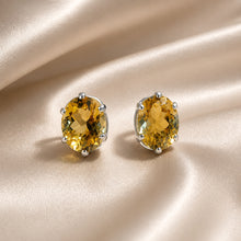 Load image into Gallery viewer, A pair of oval-cut yellow citrine gemstone stud earrings in a silver four-prong setting, displayed on a draped beige satin fabric background.

