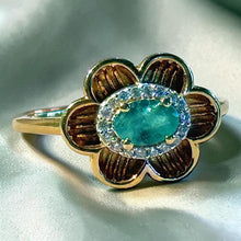 Load image into Gallery viewer, A gold flower-shaped ring featuring a central oval emerald gemstone surrounded by a halo of small clear stones, with textured gold petals.
