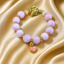 Load image into Gallery viewer, A beaded bracelet with light purple/lavender round beads, gold-toned spacer details, a gold toggle clasp, and a dangling heart charm with pink glitter, displayed on draped champagne satin fabric.
