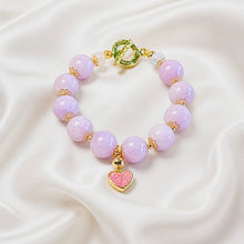 Load image into Gallery viewer, A beaded bracelet with light purple lavender beads, gold spacer rings, a gold clasp with a green accent, and a dangling pink heart charm with gold trim, displayed on a white satin fabric background.
