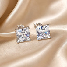 Load image into Gallery viewer, A pair of princess-cut clear cubic zirconia stud earrings in a silver setting, displayed on a draped beige satin fabric background.
