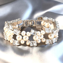 Load image into Gallery viewer, A silver-tone double-strand bracelet featuring white faux pearls and clear square-cut rhinestones, displayed on a shiny silver satin fabric background.

