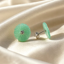 Load image into Gallery viewer, A pair of round, flower-shaped light green jadeite stud earrings with a silver-toned four-petal center, displayed on a draped cream satin fabric.
