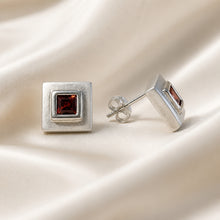 Load image into Gallery viewer, A pair of silver square stud earrings with a central square-cut dark red gemstone, displayed on a draped cream satin fabric. One earring is shown from the front, and the other is angled to show the side profile and butterfly back.
