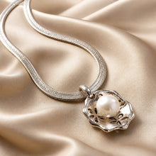 Load image into Gallery viewer, A silver snake chain necklace with a filigree pendant holding a single white pearl, displayed on draped champagne satin fabric.
