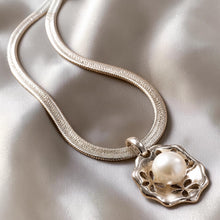 Load image into Gallery viewer, A silver-toned snake chain necklace with a decorative pendant featuring a white pearl, displayed on a draped satin fabric background.
