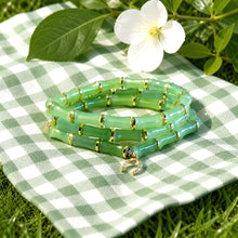 Load image into Gallery viewer, A set of green beaded stretch bracelets with gold spacer rings, coiled on a green and white checkered blanket outdoors with grass and a white flower in the background.
